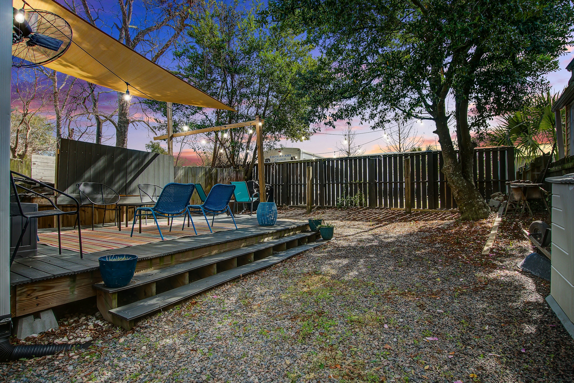 Backyard at dusk: wooden deck with two blue lounge chairs, beige shade sail, dark wood privacy fence, gravel yard, and warm string lights.