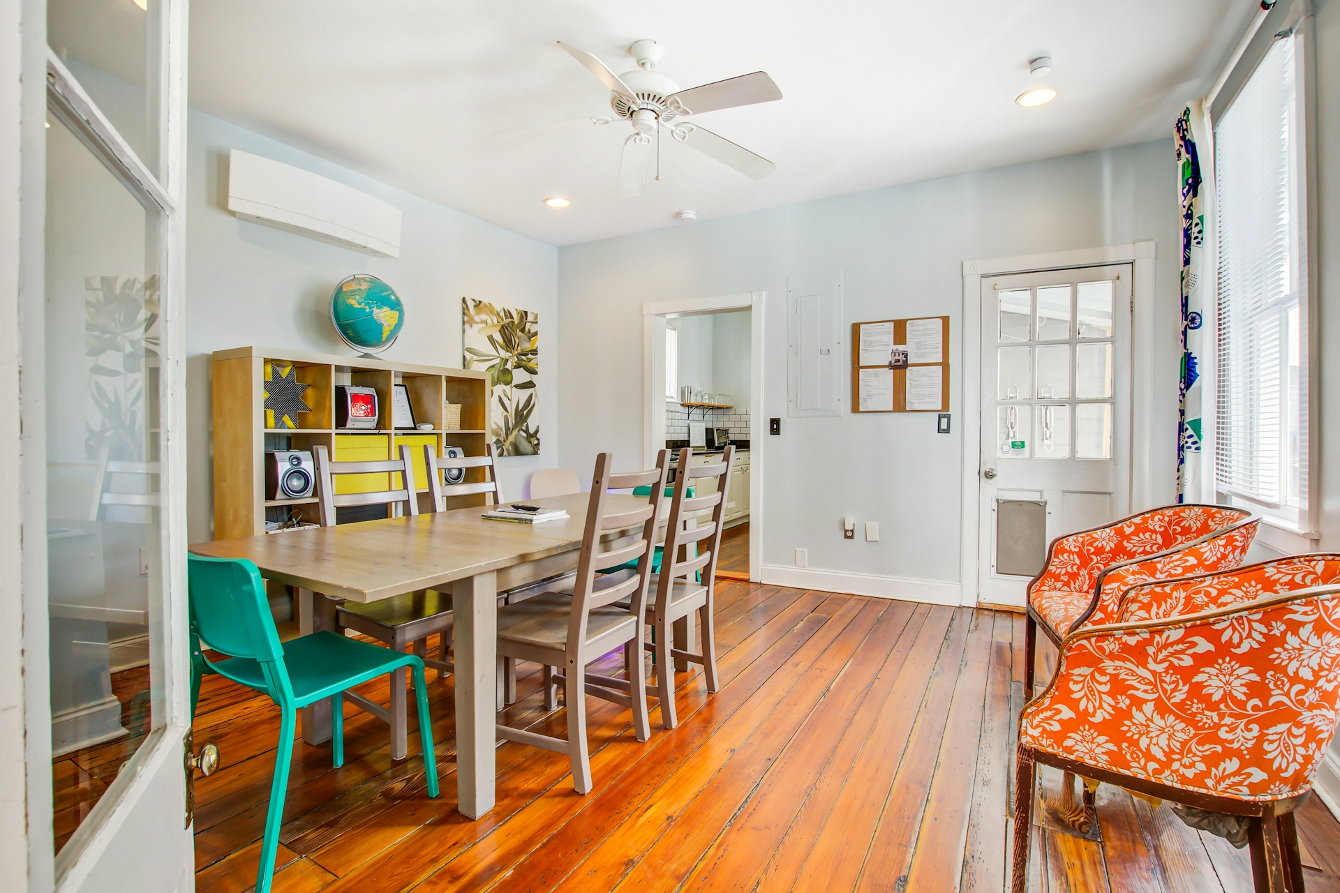 Dining area with long wooden table, mixed chairs including teal accents and orange floral armchairs, cube shelving with globe, wide-plank pine floors, and ceiling fan.