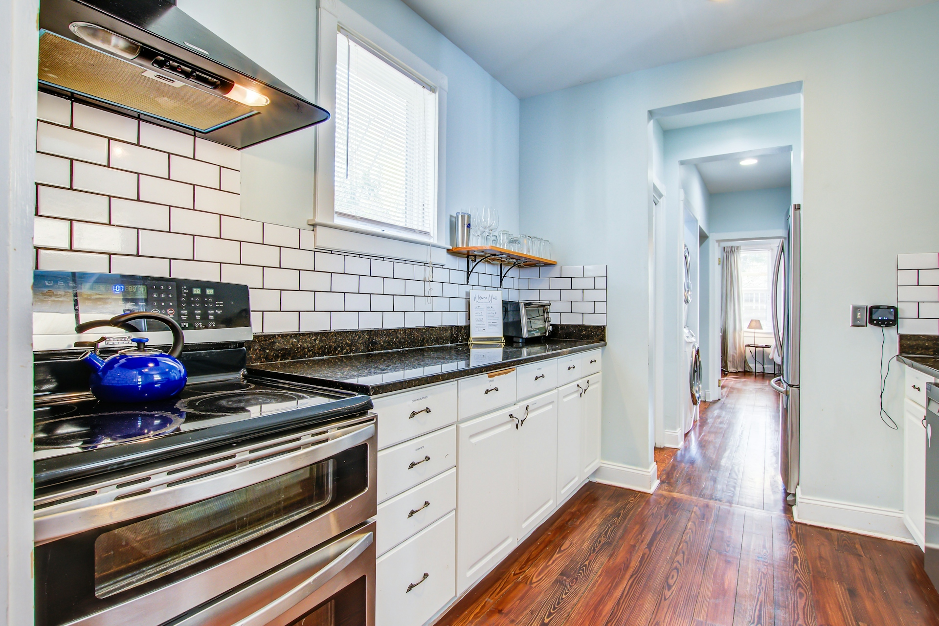 Kitchen stove area: stainless double oven and range with blue kettle, range hood, white subway tile, dark granite counters, stemware shelf, view toward laundry hallway.