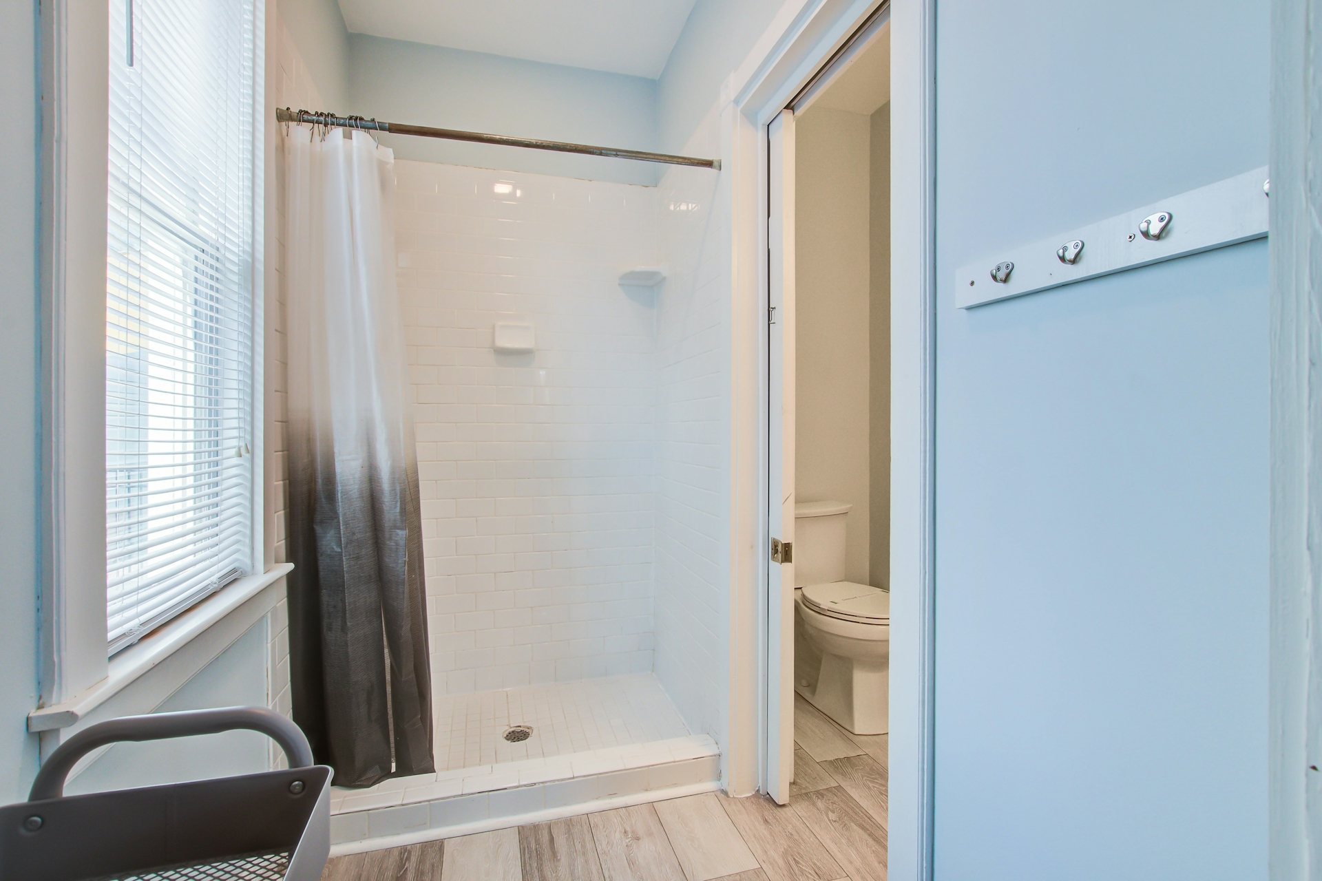 Bathroom with large walk-in subway-tile shower, gray-and-white ombre curtain, window with blinds, and adjoining toilet alcove.