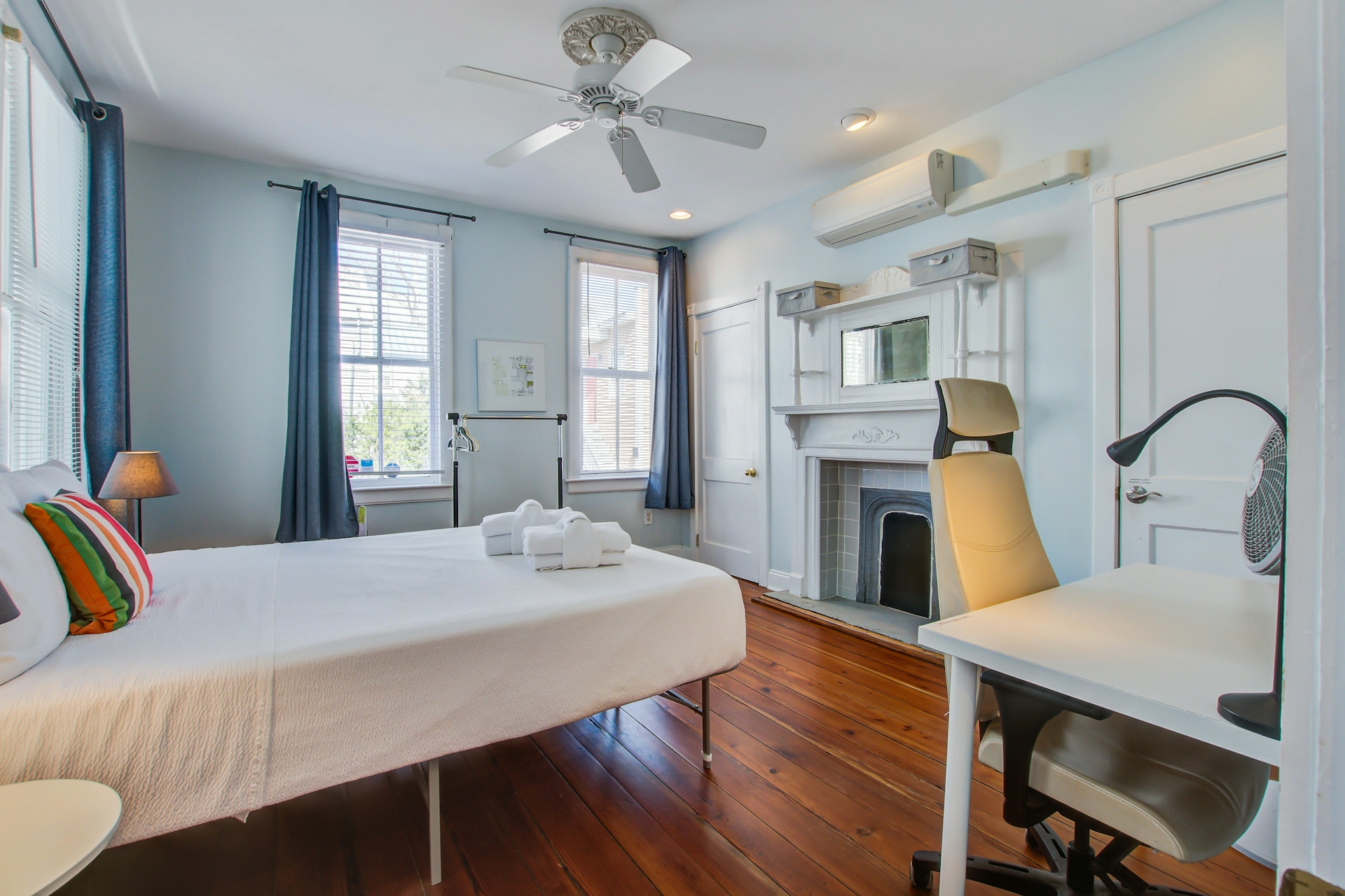 Bedroom with decorative white fireplace mantel, queen bed with striped pillow, desk with office chair and fan, blue curtains, and mini-split AC.