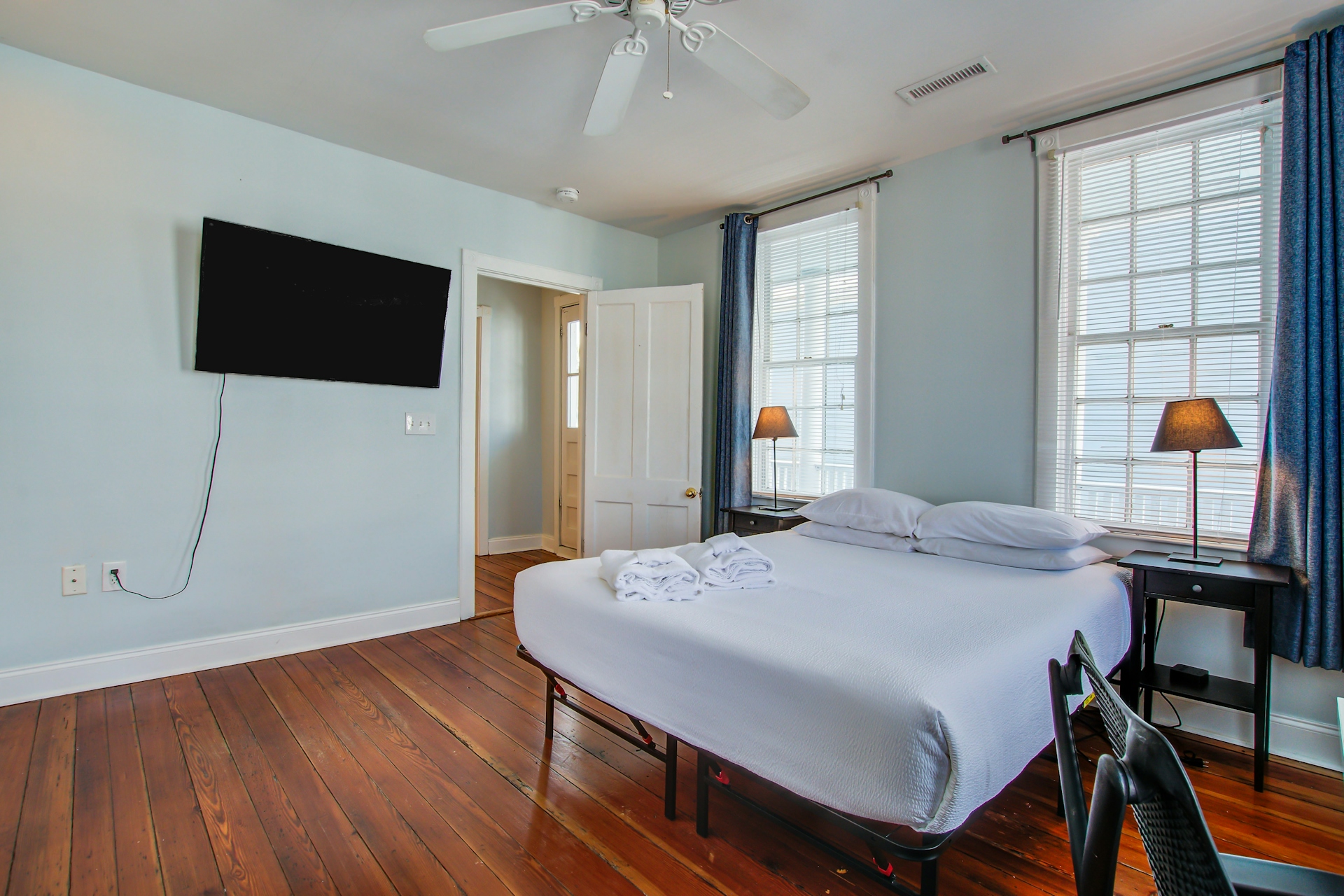 Bedroom with wall-mounted TV, queen bed, two black bedside lamps, two tall windows with blue curtains, and doorway to porch or balcony.