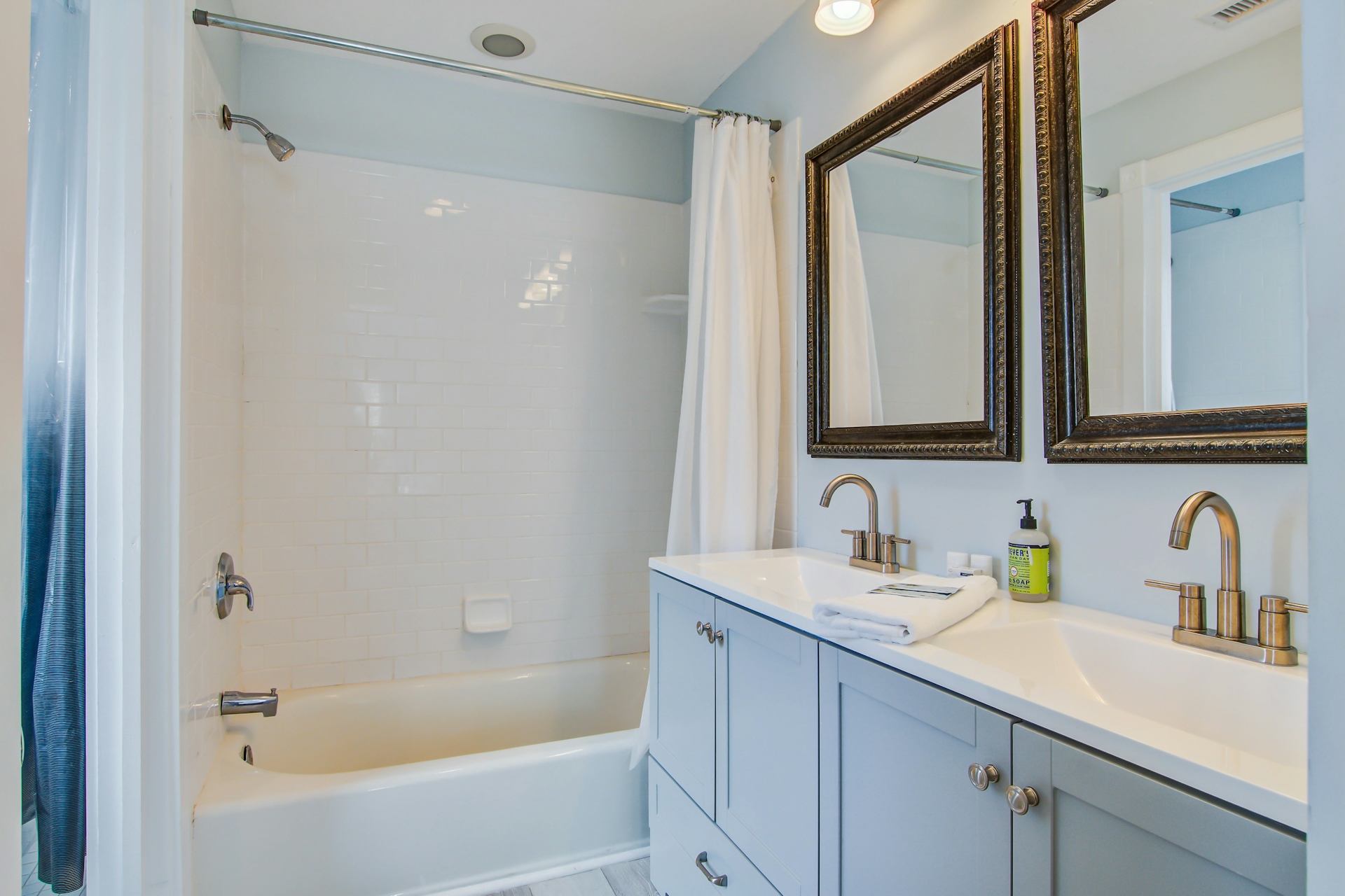 Bathroom with gray double-sink vanity, two ornate framed mirrors, white tub-shower with subway tile and shower curtain.