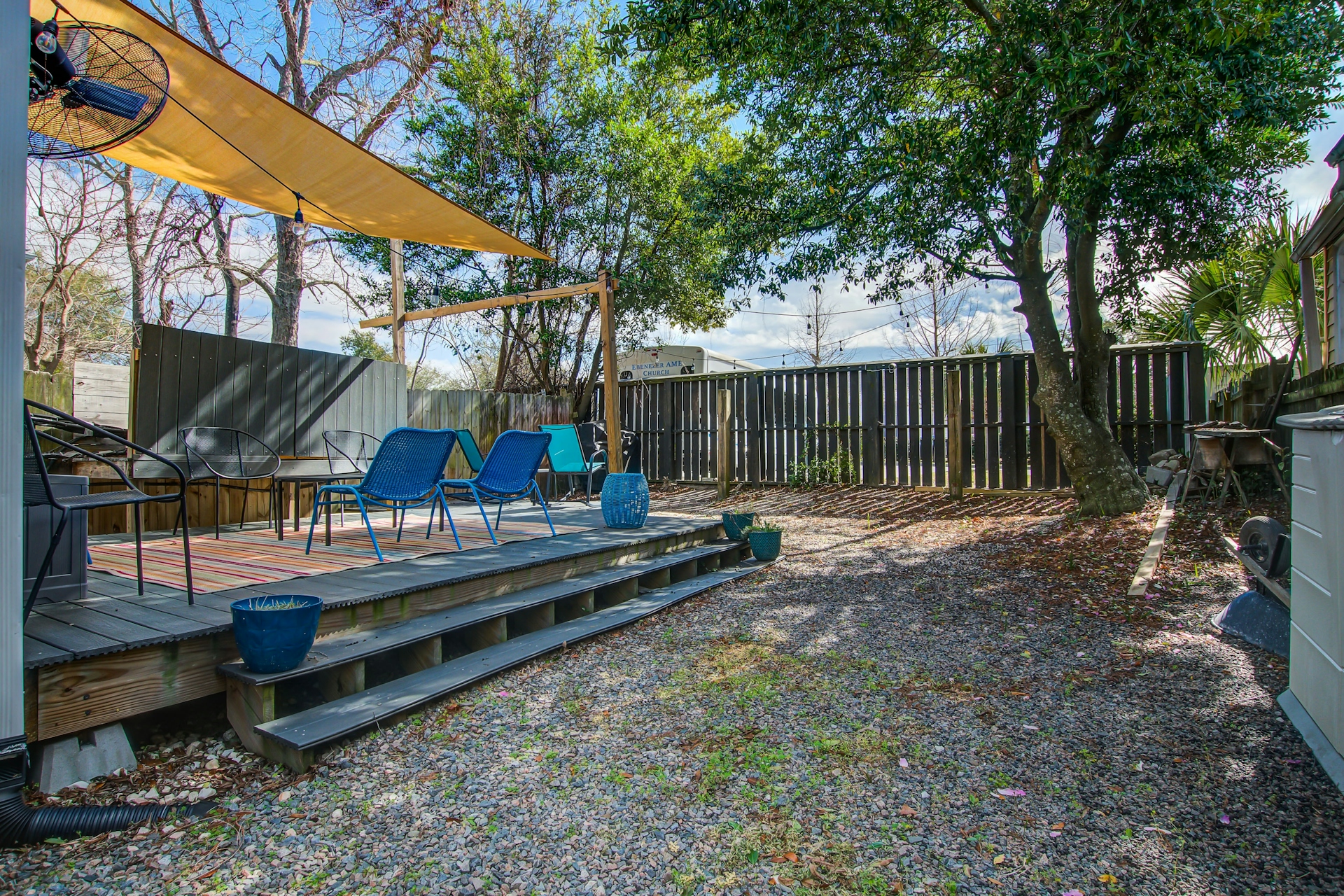 Daytime backyard deck with two blue lounge chairs, shade sail, gravel yard, large oak tree, and dark wood privacy fence.