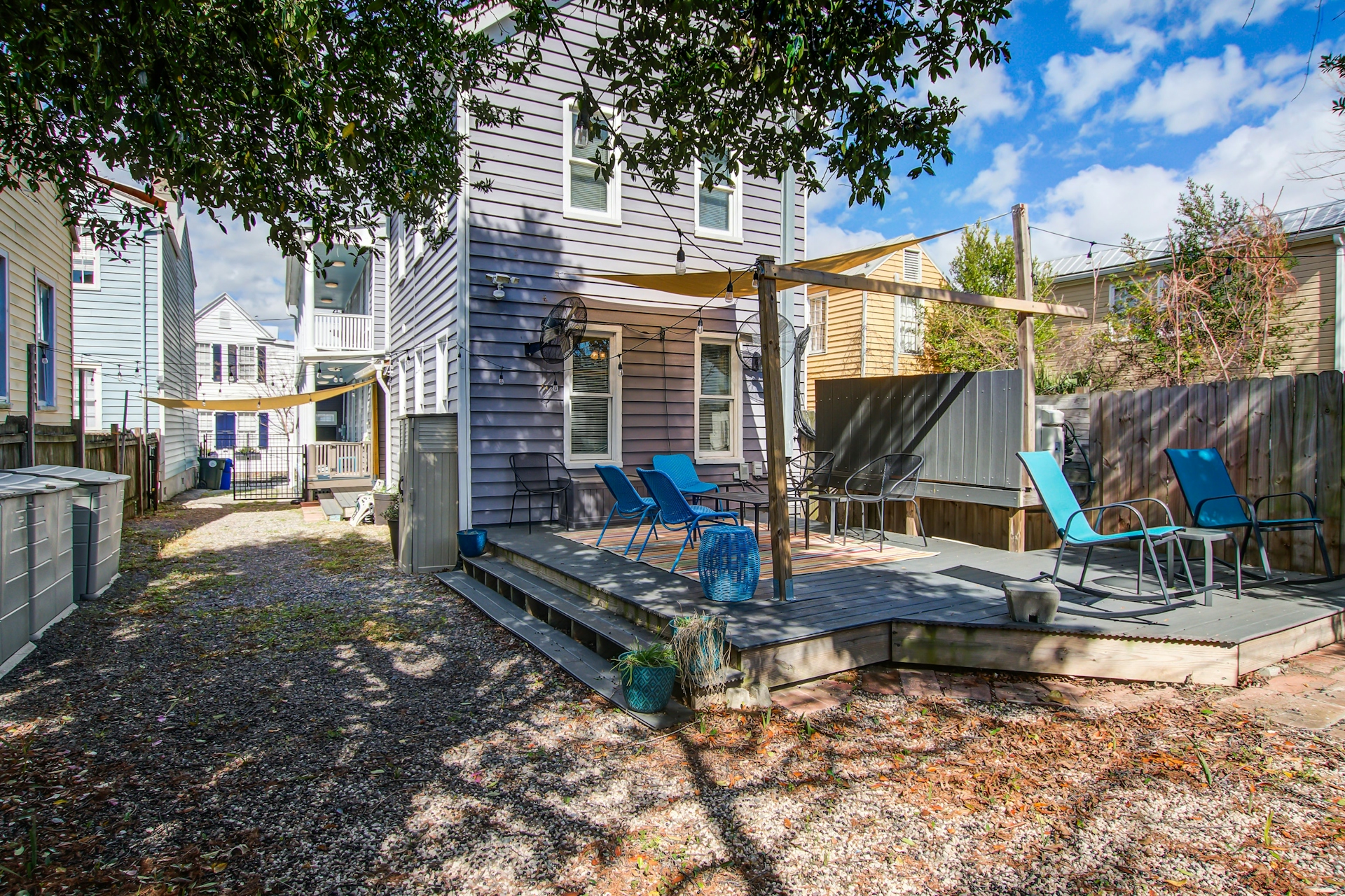 Rear exterior of the house: lavender siding, multiple windows, shade sail over deck, blue lounge chairs, and gravel yard.